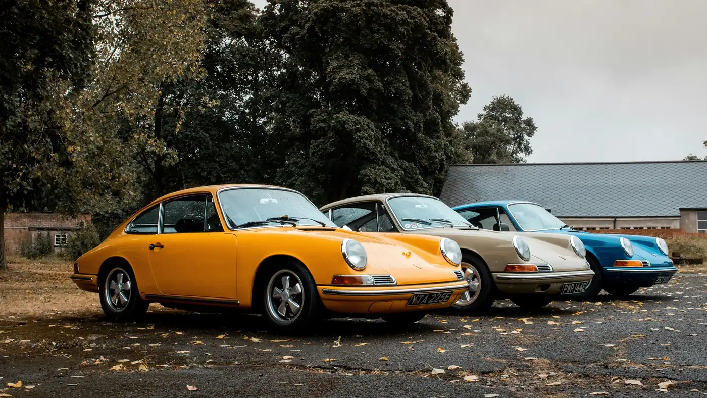 Tres Porsches clásicos en fila (amarillo, crema y azul) en un entorno natural